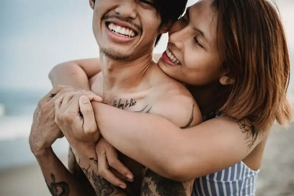 A young couple embraces playfully on the beach, showcasing tattoos and joyful smiles.