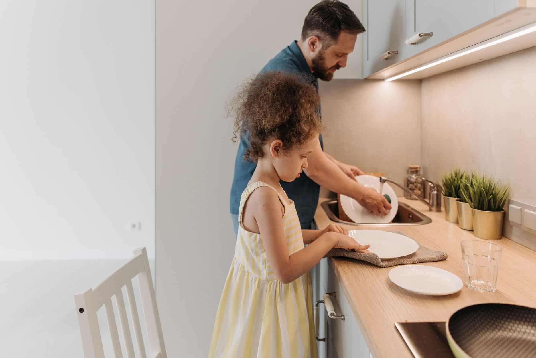 Young girl drying a dinner plate at the kitchen counter while an adult washes dishes at the sink