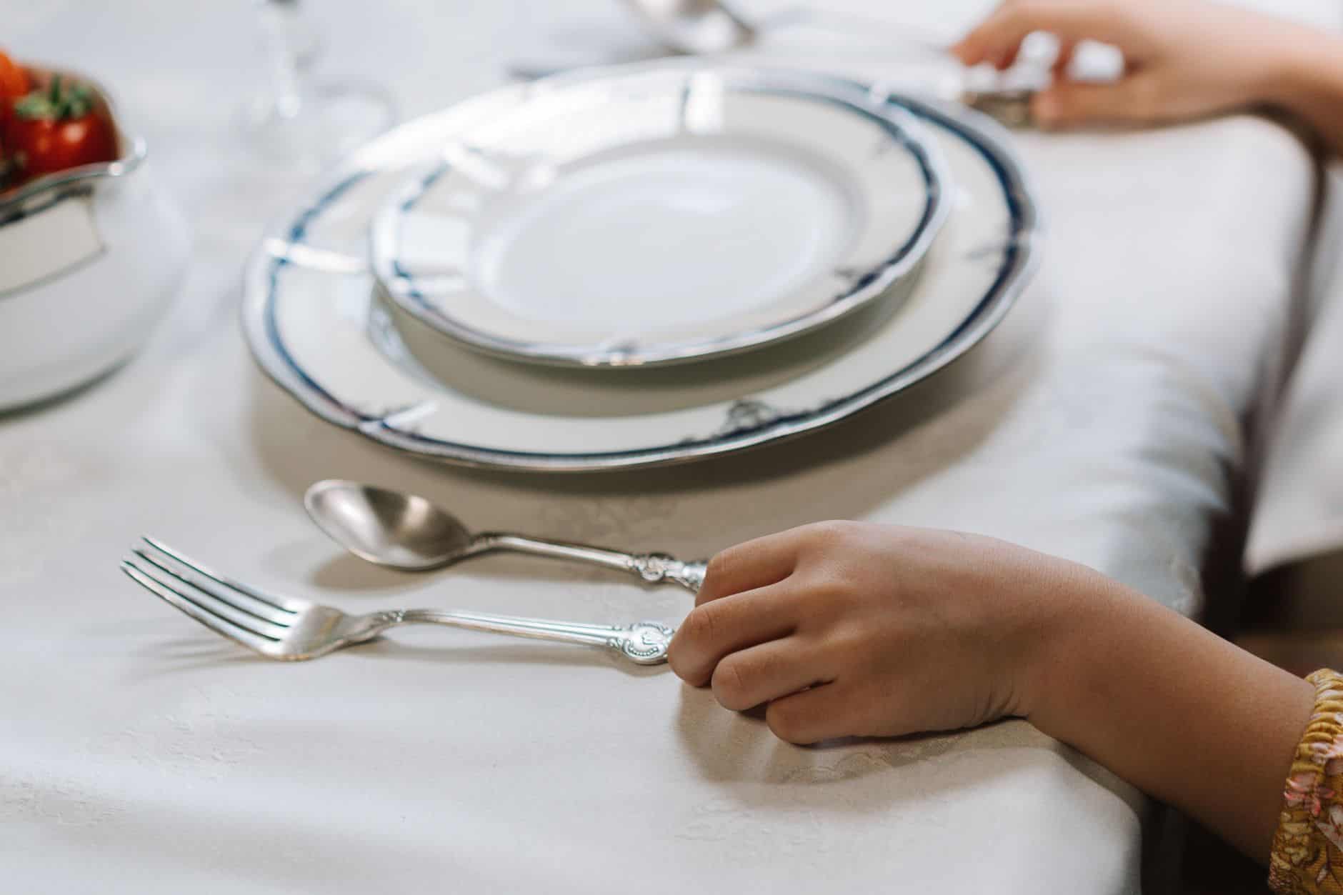 Child's hands resting beside an empty plate and silverware at a set dinner table
