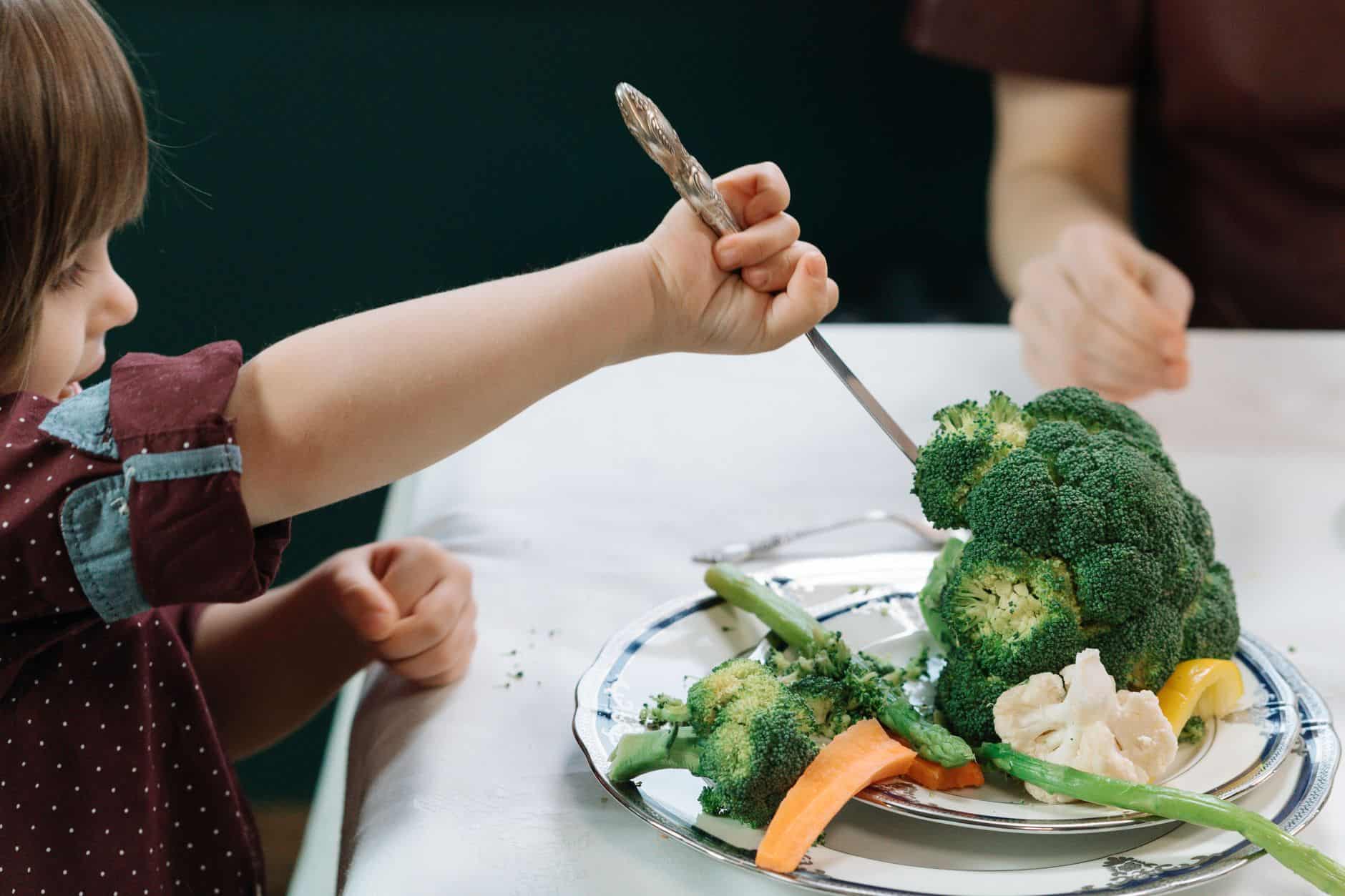 Young child using a fork to try broccoli and other vegetables on a dinner plate