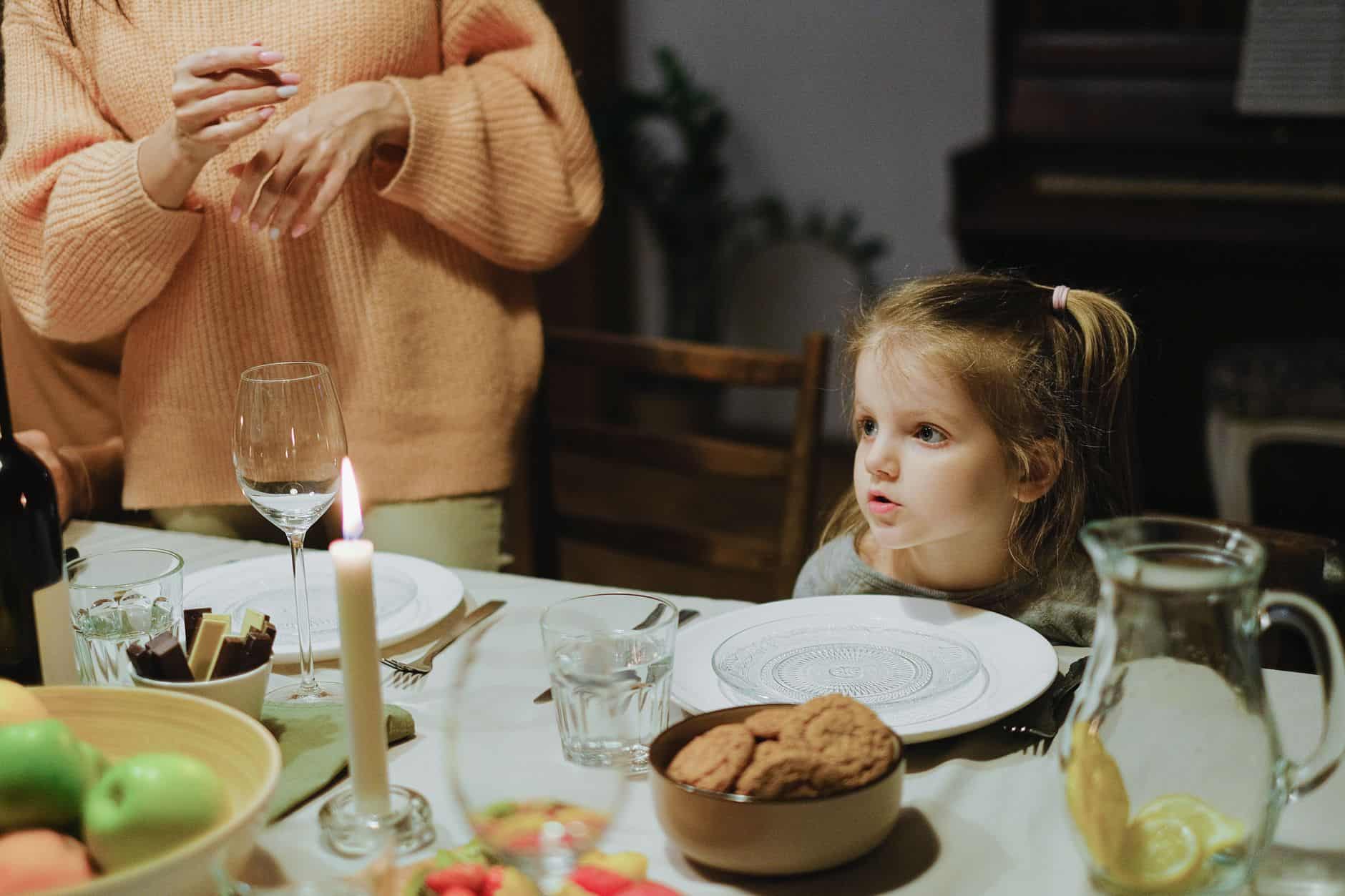 Young girl seated at a set dinner table with an empty plate, waiting while an adult still stands beside the chair