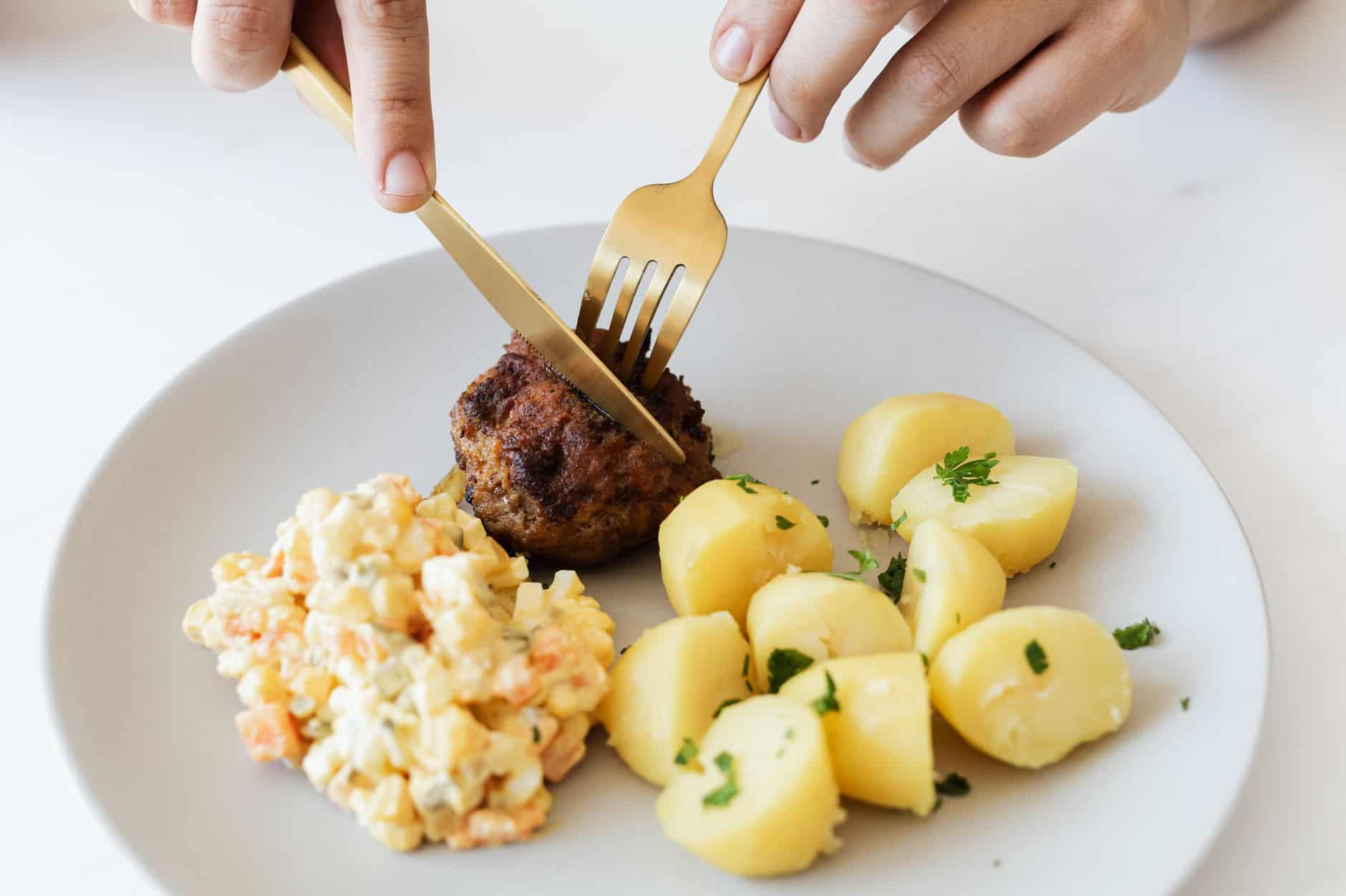 Hands using a knife and fork to cut a bite of meat on a plate with potatoes and salad