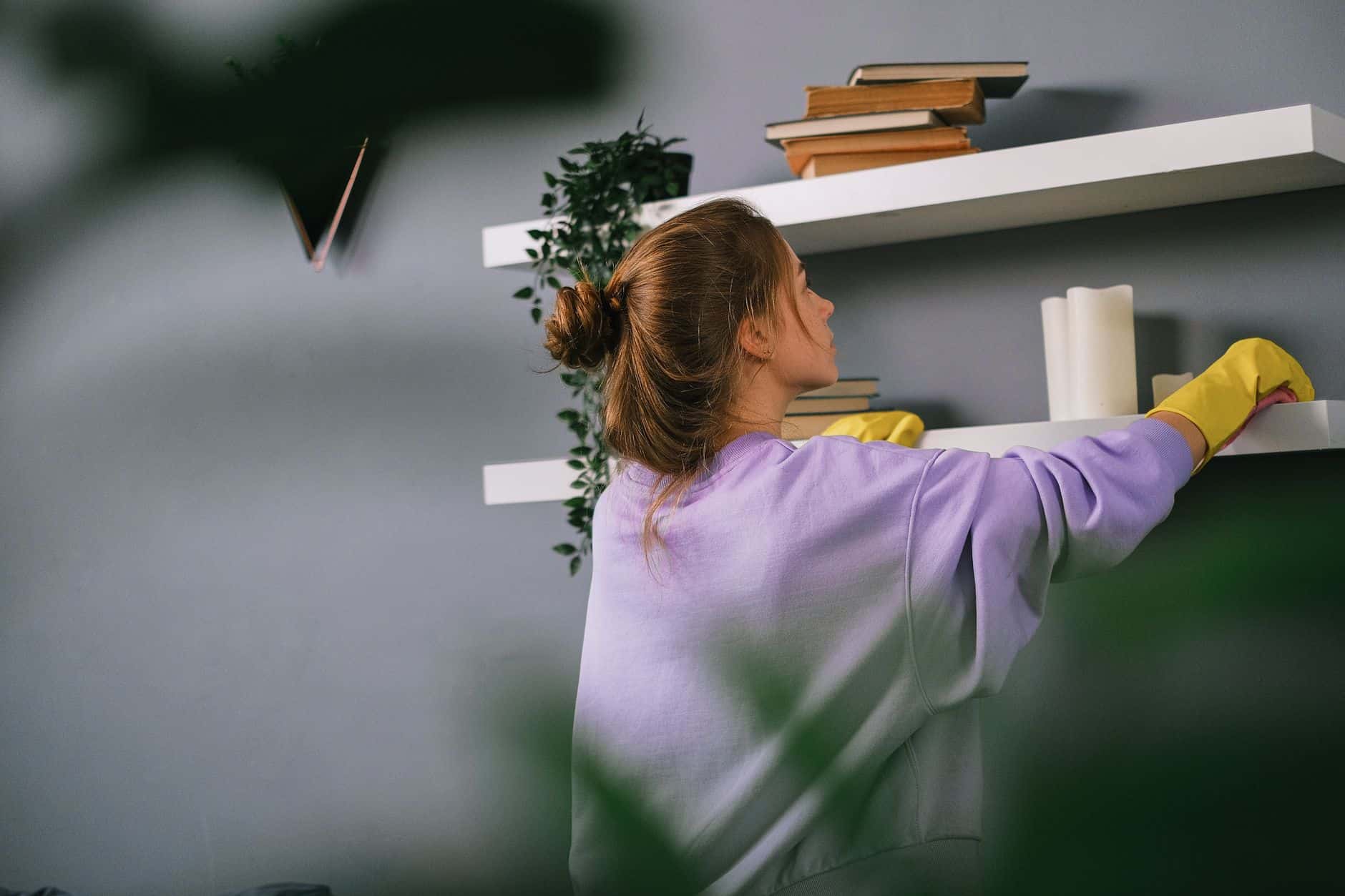 Woman in yellow gloves wiping a high white shelf from the top down