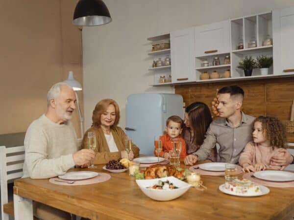 Grandparents, parents, and two children seated around a kitchen table with empty plates and a roast in the center