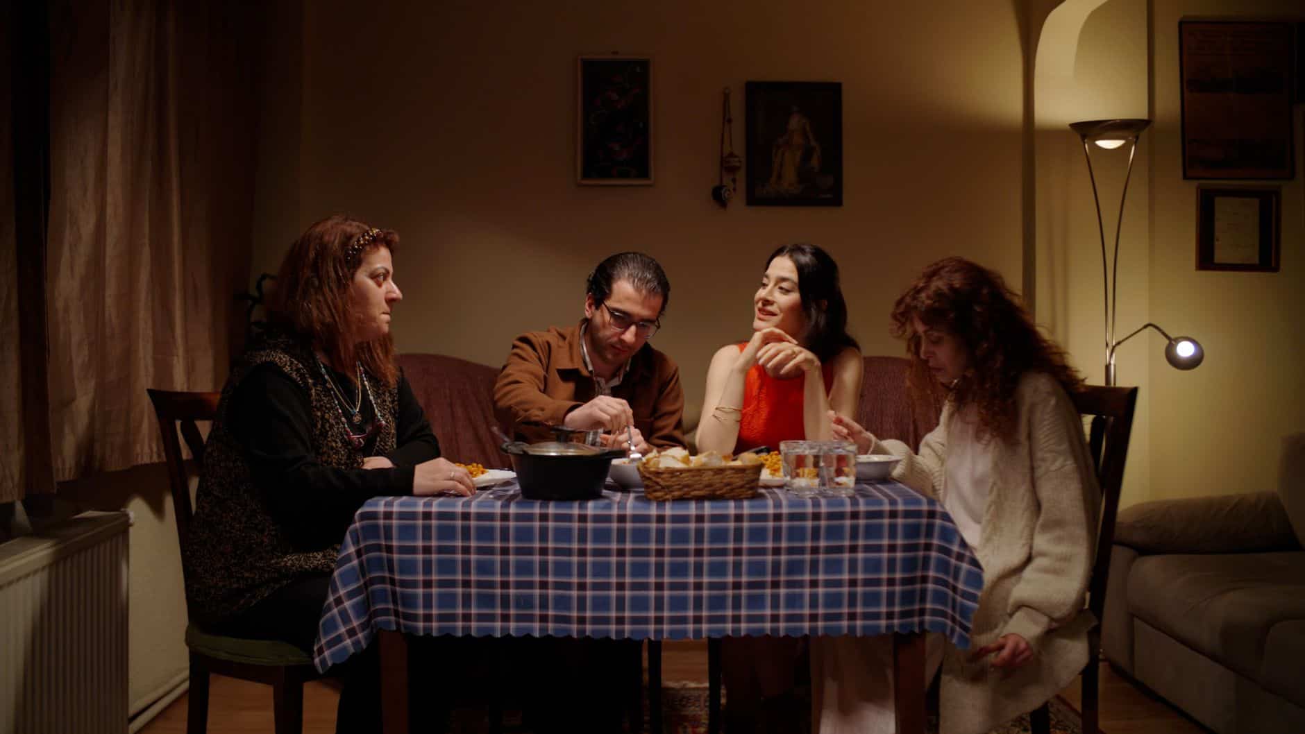 Four people seated at a small kitchen table for dinner, sitting up with their hands near their plates