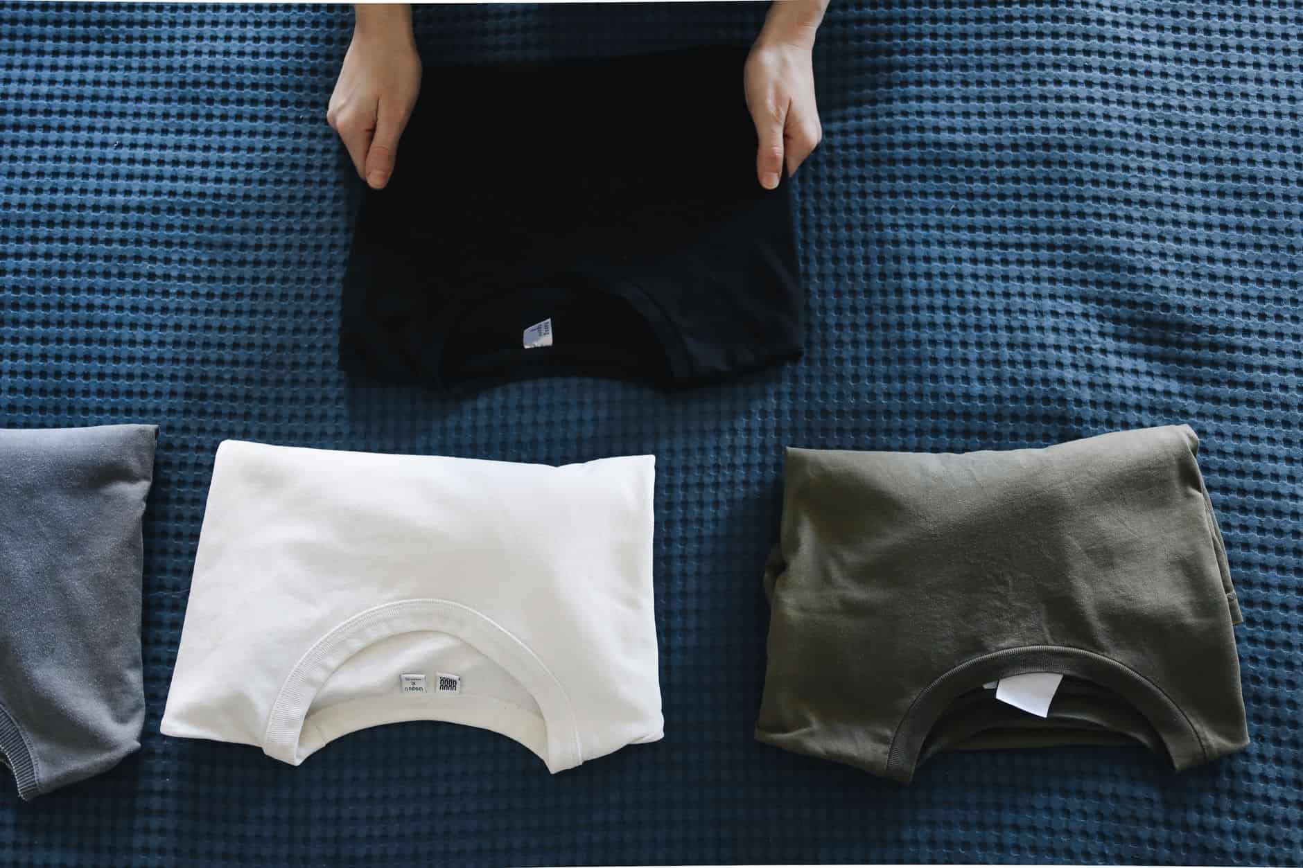 Hands folding a black t-shirt next to other folded shirts on a bed