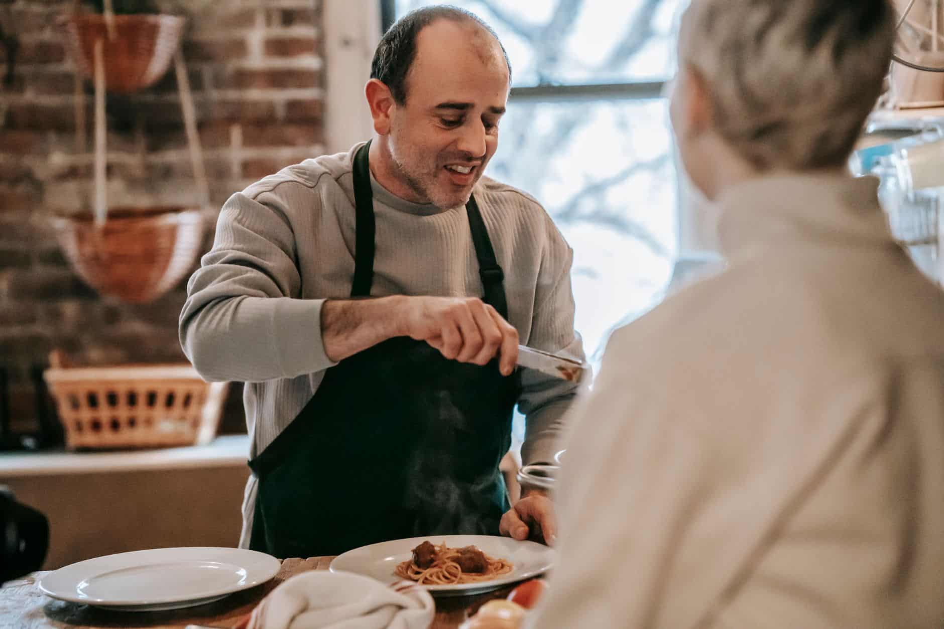 Smiling man in an apron serving a plate of spaghetti and meatballs at the kitchen table