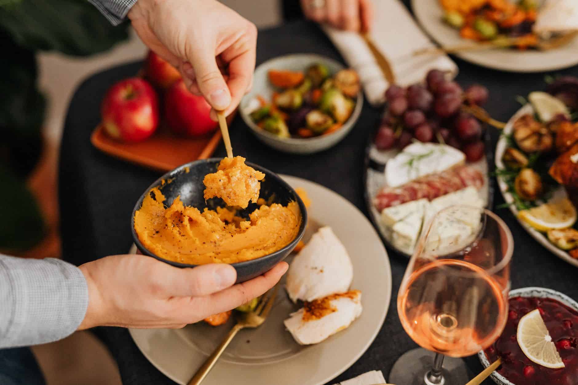 Hands scooping mashed sweet potatoes from a bowl onto a dinner plate with a serving spoon