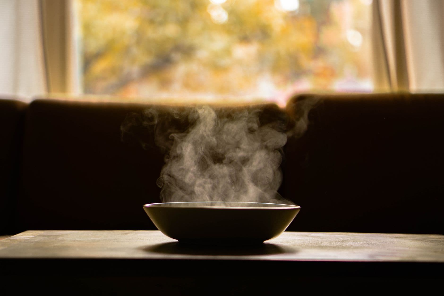 A steaming bowl of hot soup sitting on a table in front of a window