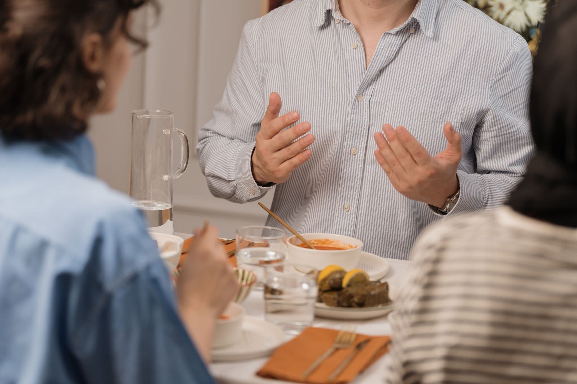 People at a dinner table listening while one person talks with their hands, plates and glasses in front of them