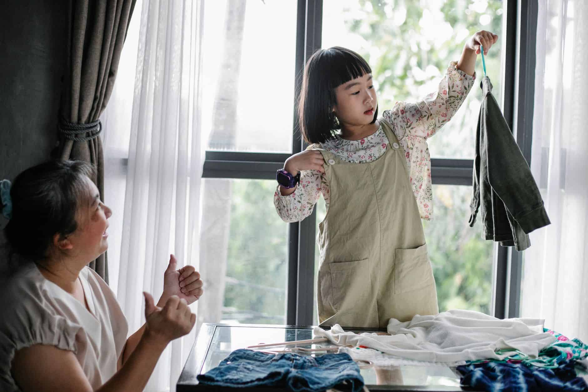 Grandmother watching her granddaughter hold up clothes beside a pile of laundry
